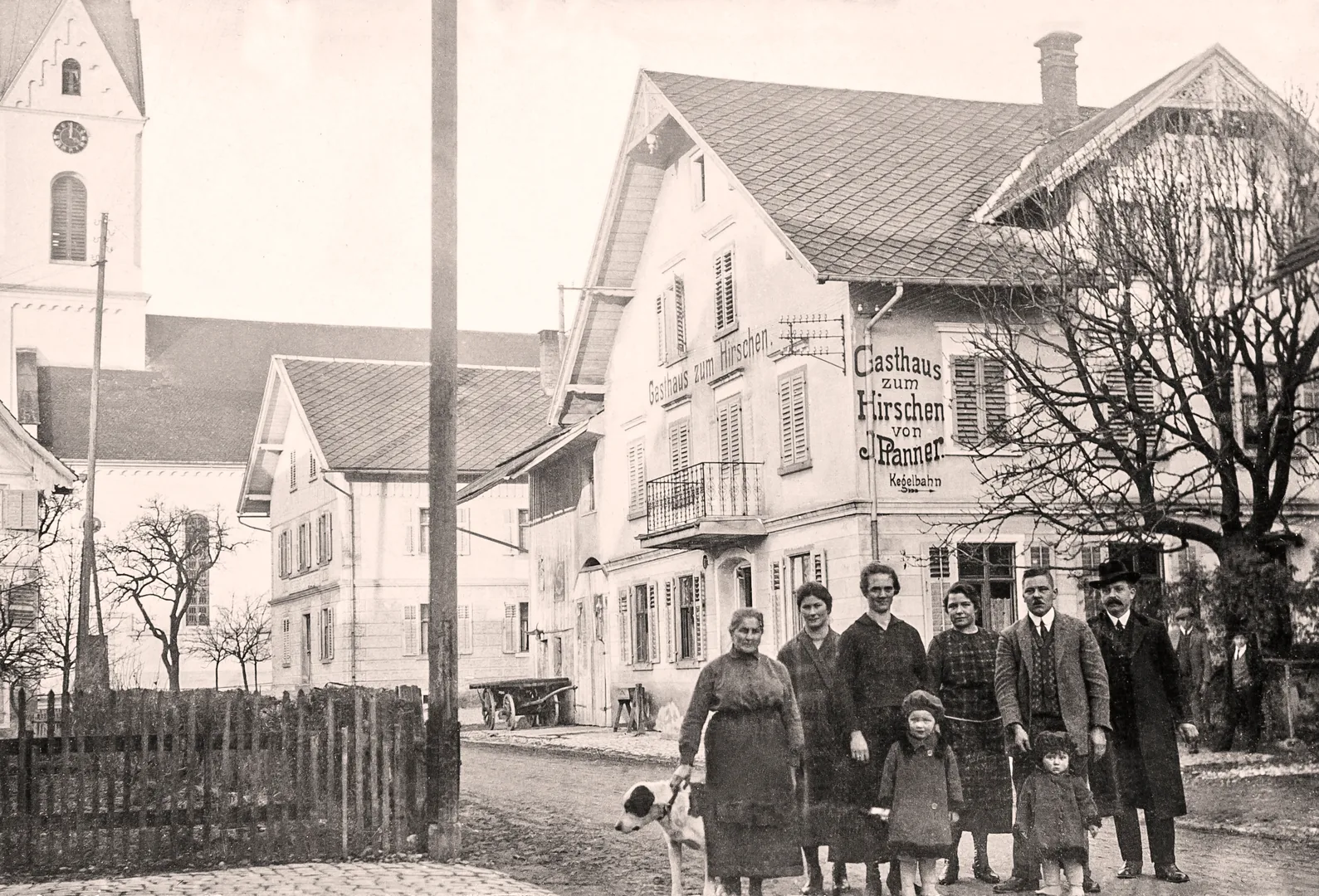 Historische Schwarz-Weiß-Aufnahme: Die Familie Pfanner mit Kindern und Hund steht vor dem Gasthaus zum Hirschen in Lauterach, im Hintergrund die Dorfkirche.