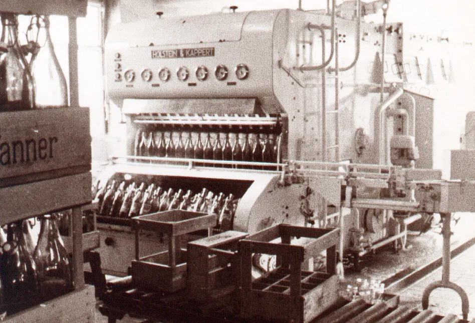 Historische Aufnahme einer Flaschenreinigungs- und Abfüllmaschine der Marke Holstein & Kappert mit Bügelflaschen und Pfanner-Holzkisten.