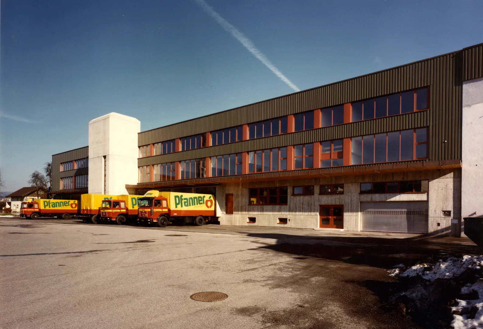 Das Firmengebäude am Stammsitz Lauterach mit drei gelb-roten Pfanner-Lastwagen davor unter blauem Himmel.