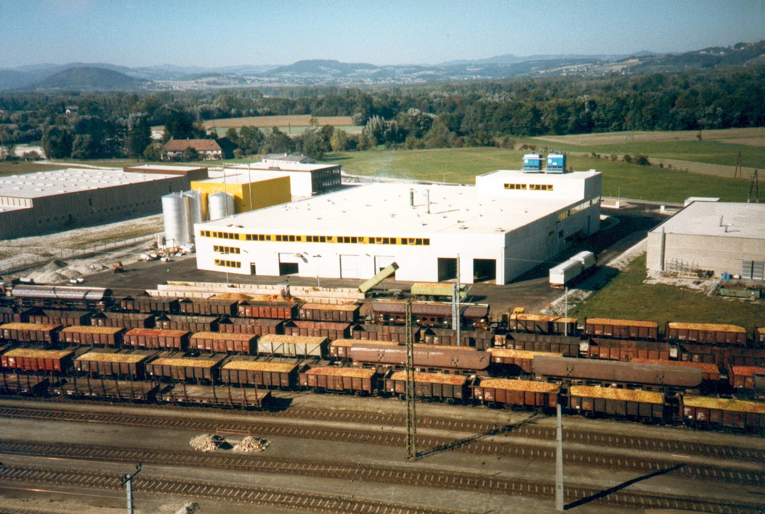 Luftaufnahme des Werks in Enns mit langen Reihen voll beladener Apfel-Eisenbahnwaggons davor, im Hintergrund Felder und Hügel.