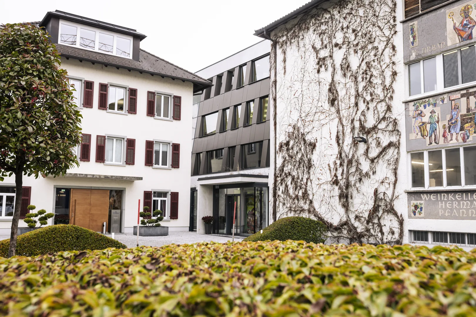 Der heutige Pfanner-Firmensitz in Lauterach: ein historisches Gebäude mit roten Fensterläden neben einem modernen Anbau, an der efeubewachsenen Fassade Mosaike und der Schriftzug Weinkeller Hermann Pfanner.